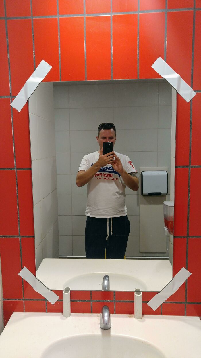 Hilariously bad DIY: A bathroom mirror taped to a red tiled wall, reflecting a man taking a selfie.