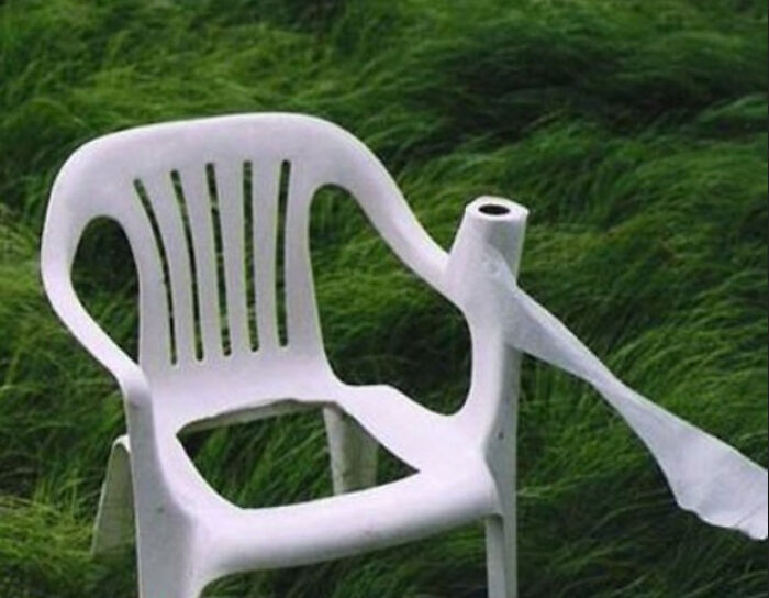 Hilariously bad DIY: A white plastic chair with a toilet paper roll taped to its armrest, on green grass.