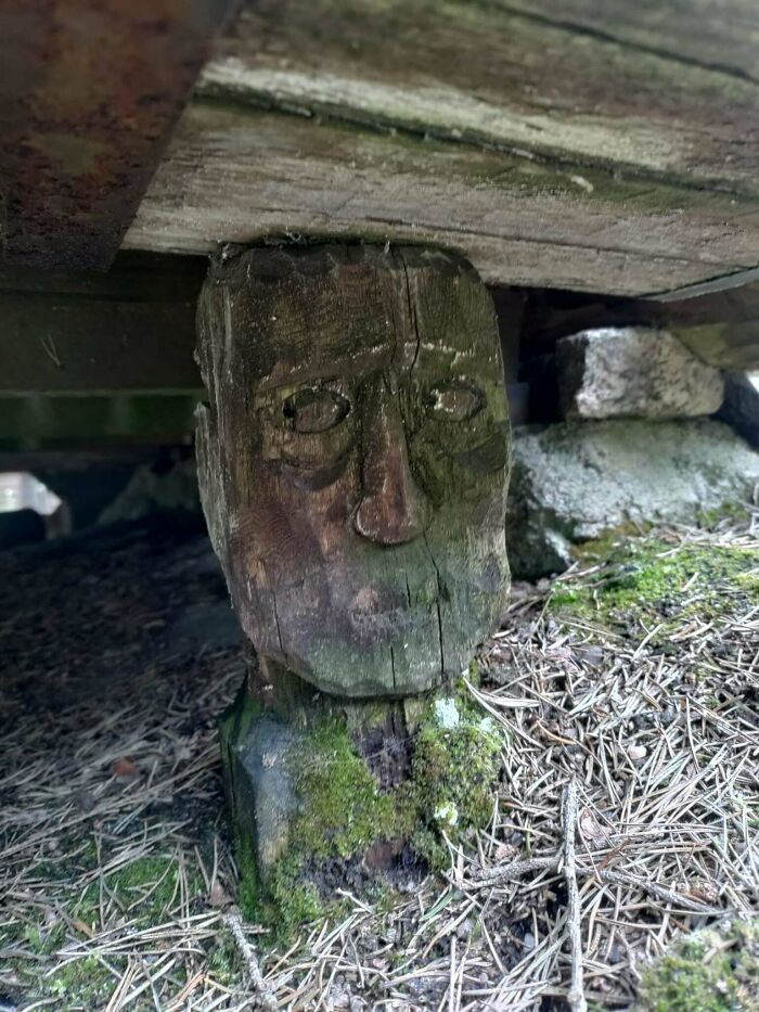 A weathered wooden carving of a face, seemingly supporting a structure, an unexpected moment in nature.