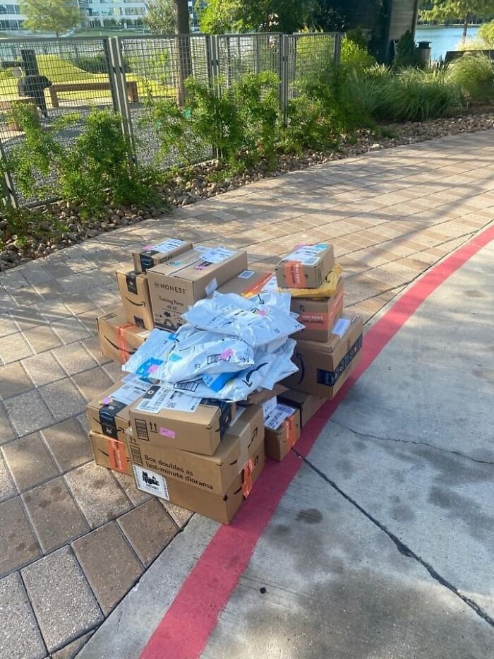 A large pile of Amazon packages and envelopes stacked on a sidewalk, a mildly infuriating obstacle for delivery.