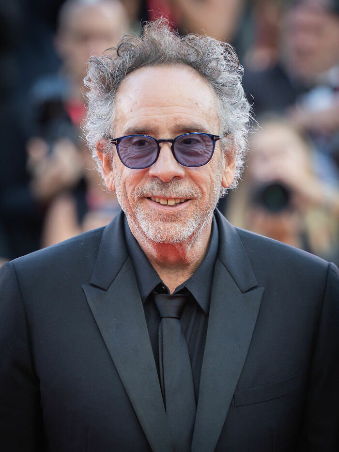 A close-up of Tim Burton, wearing sunglasses and a black suit, smiling at an event, highlighting his career.