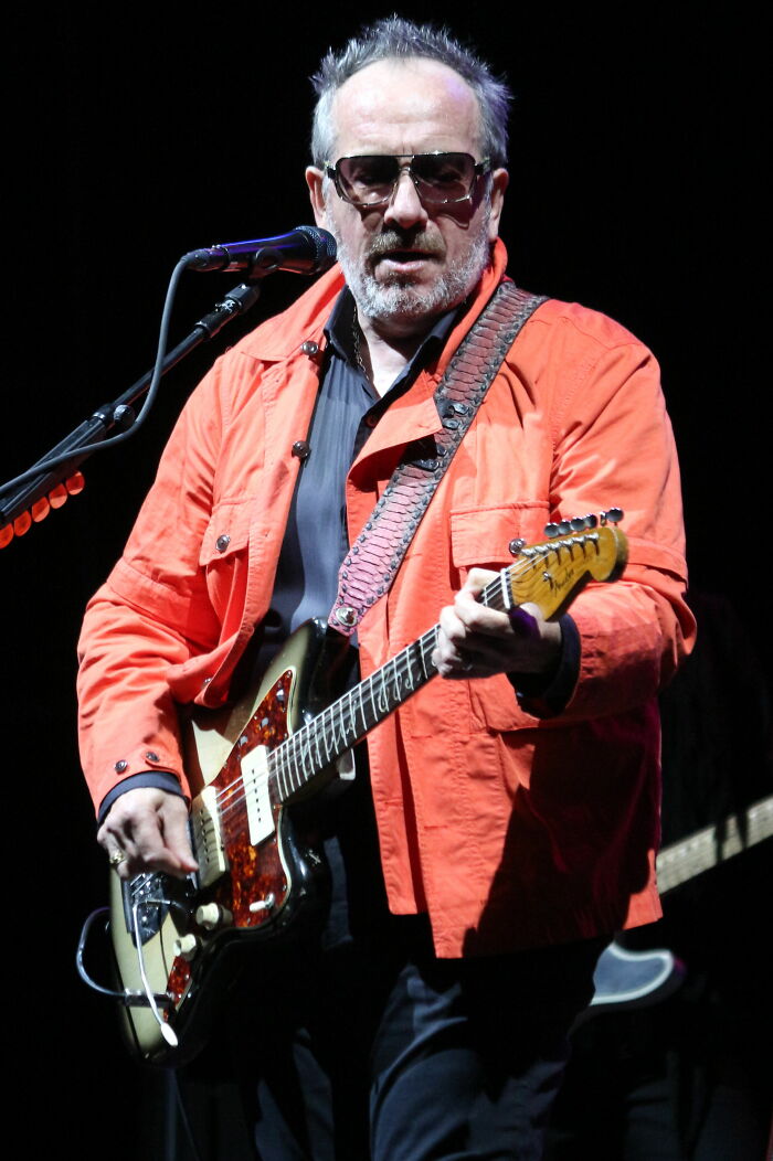 Elvis Costello playing electric guitar on stage, wearing sunglasses and an orange jacket, highlighting his career.