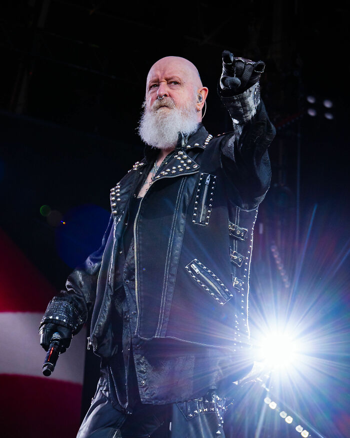 Rob Halford, a bald man with a long white beard, on stage in a studded leather jacket holding a microphone.