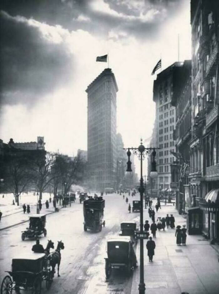 An iconic photo from American history: the Flatiron Building in a bustling city street with horse-drawn carriages and pedestrians.