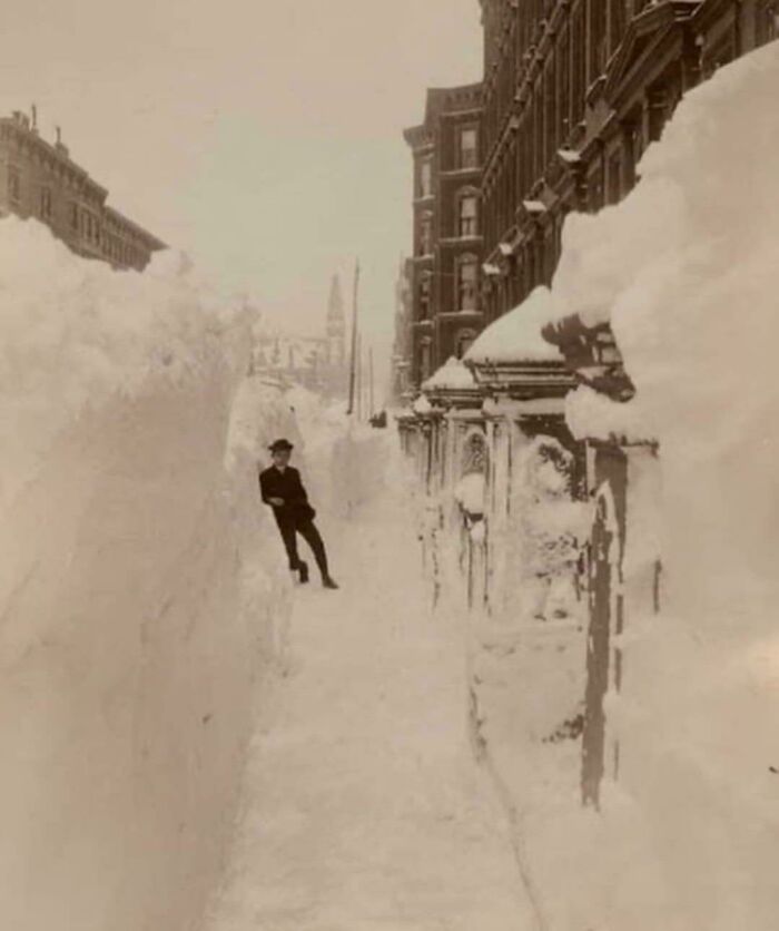 A man stands dwarfed by towering snowdrifts, a fascinating photo from American history during a massive blizzard.