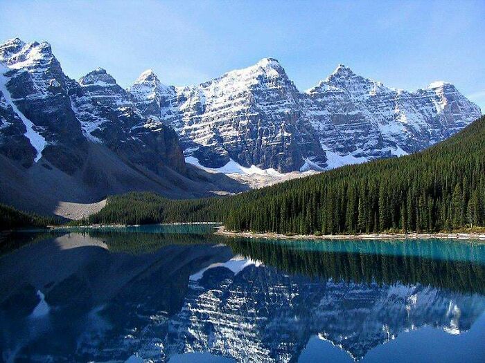 A stunning landscape photo from American history with snow-capped mountains reflecting in a clear blue lake, surrounded by dense pine forests.