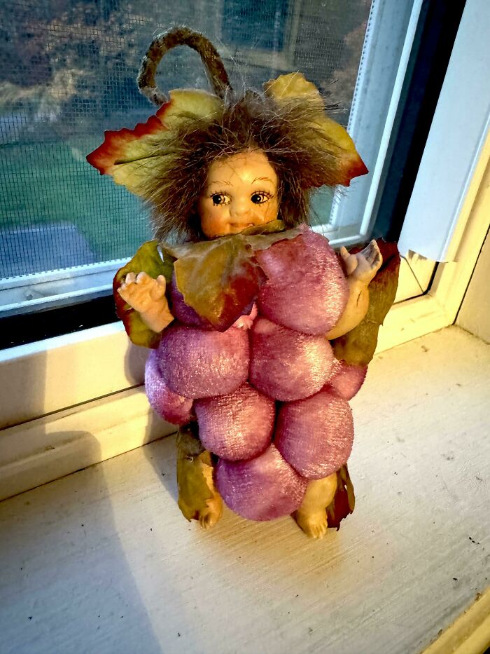 A small doll dressed as a bunch of grapes, with a doll face and brown hair, representing things people lost hope to find.