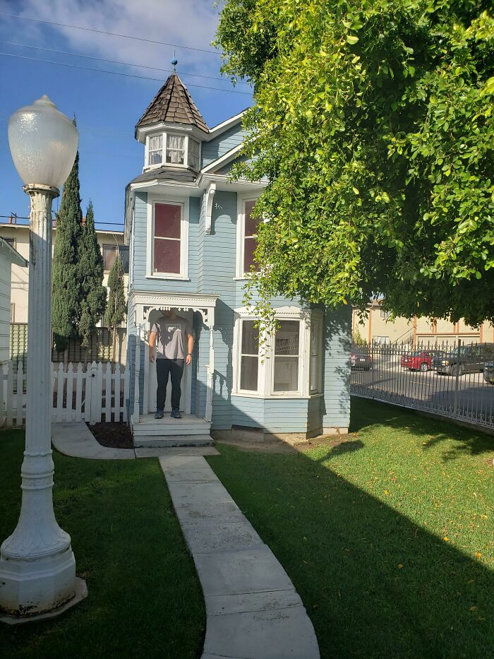 A person standing under a small porch of a miniature-looking house, raising questions about the Airbnb listing scale.