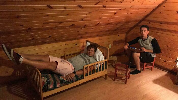 A man lies on a tiny bed in an Airbnb listing, with another man sitting on a small chair, highlighting unexpected accommodations.