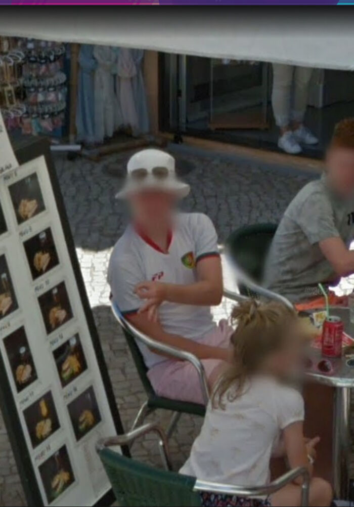 A blurred man in a white hat and shirt sitting at an outdoor cafe table, an odd sight on Google Maps Street View.