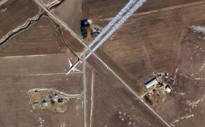 An airplane flying over fields, leaving a long contrail, as seen on Google Maps, with a road winding through the landscape.