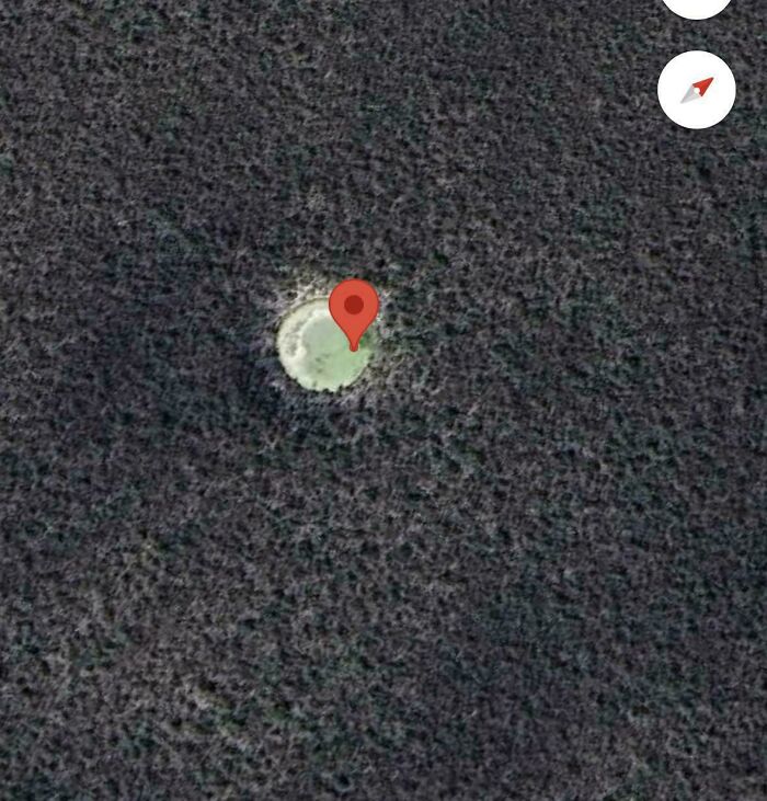 An aerial view of a small, light-colored circle in a dark, textured field on Google Maps, marked with a red pin.