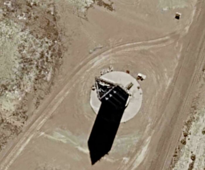 A satellite view of an odd structure on Google Maps, casting a long shadow on a barren landscape.