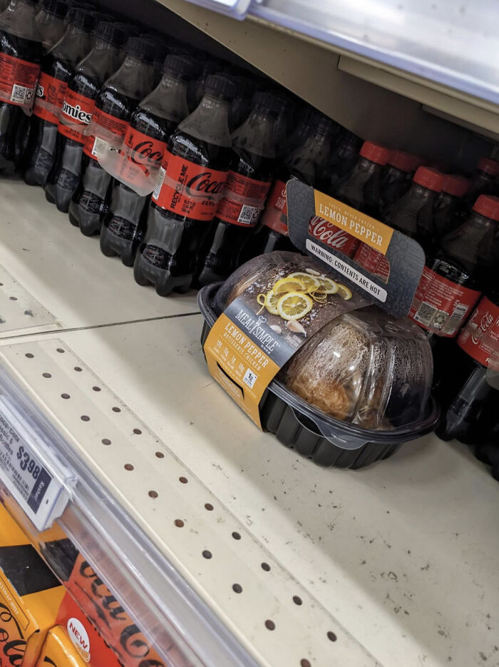 A rotisserie chicken in a plastic container with lemon pepper seasoning next to a shelf of Coca-Cola bottles. Little hope for humanity.