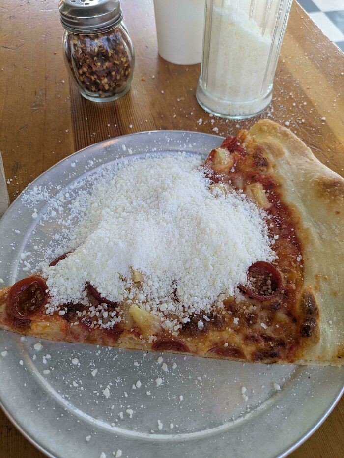 A slice of pizza heavily covered in parmesan cheese, alongside a shaker of red pepper flakes, depicting little hope for humanity.