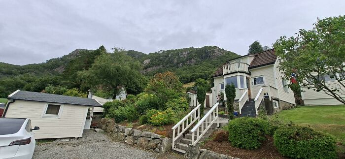 A wide shot captures a charming Airbnb listing house nestled in a green, mountainous landscape under a cloudy sky.