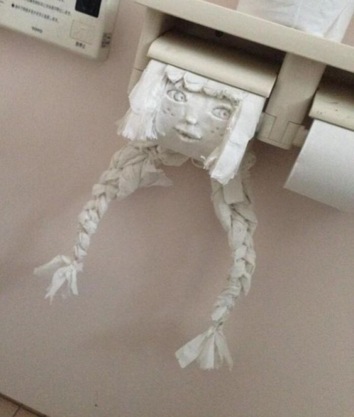 A toilet paper holder with an absolutely unhinged face and braided hair made of toilet paper rolls, a ridiculous bathroom decoration.
