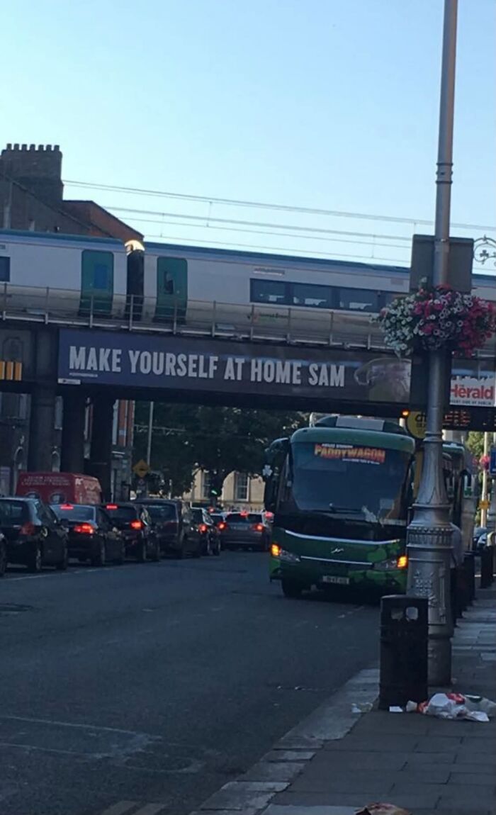 A bus and train pass under a bridge with a sign that reads Make Yourself At Home Sam, a sign from the universe.