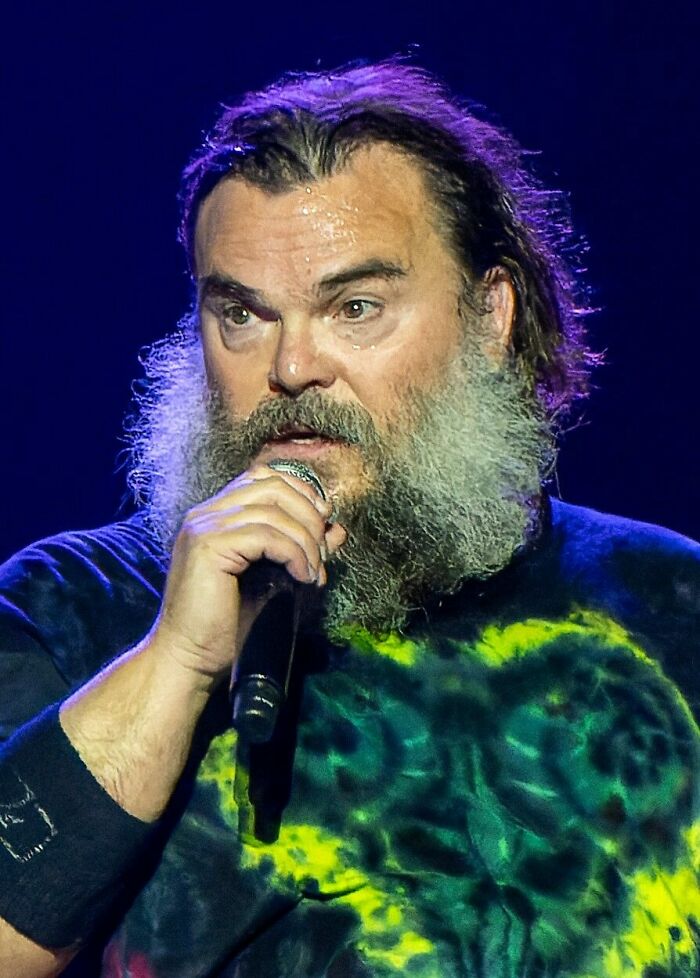 Jack Black performing on stage, wearing a tie-dye shirt, holding a microphone. His career highlights include many films.