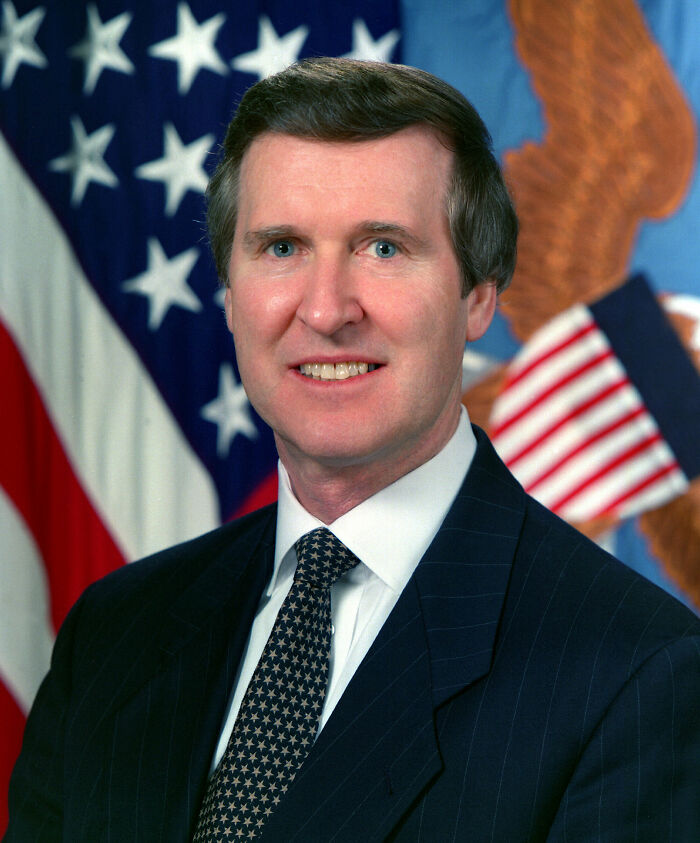 A portrait of William Cohen in a suit and tie, smiling, with the American flag in the background, showcasing his bio and career highlights.