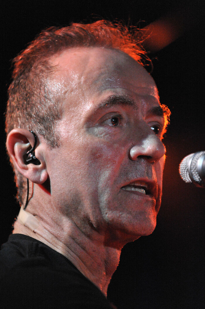 A close-up of Hugh Cornwell singing into a microphone, illuminated by red stage lighting during a performance.