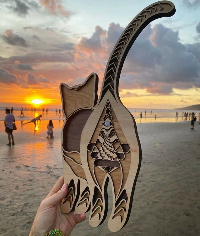 A hand holding a carved wooden cat with intricate patterns against a sunset beach scene, showcasing disturbing levels of skill.