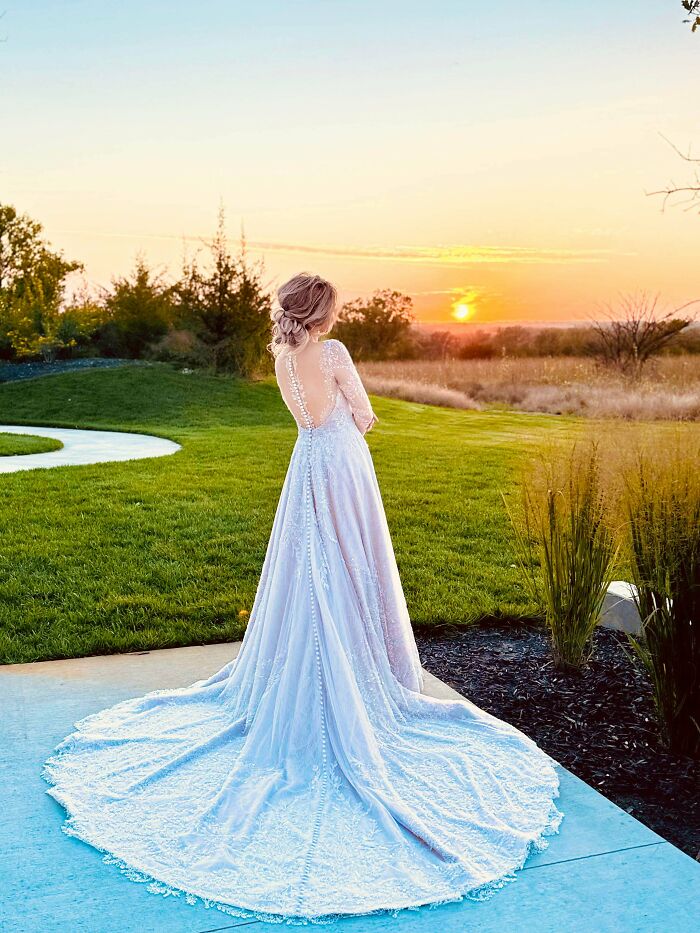 A bride in a white gown with a long train, looking at a sunset over a field, showcasing amazing crafts.