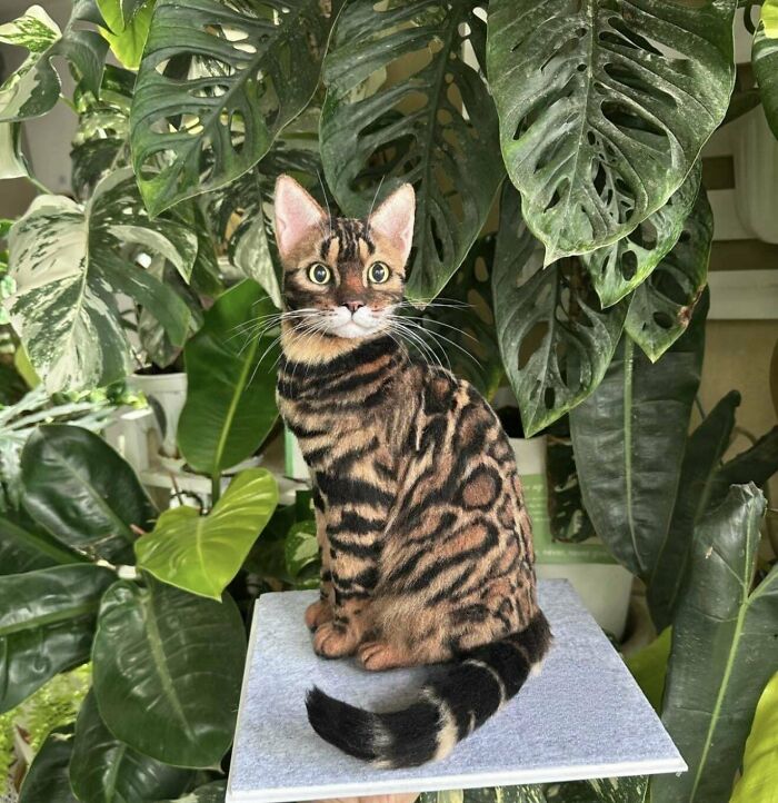 A realistic felted cat with tabby markings sitting among green plants, an example of amazing crafts.