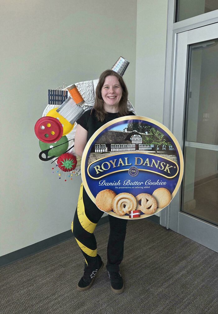 A woman in a creative sewing-themed costume, holding a giant Royal Dansk cookie tin, showcasing amazing crafts.