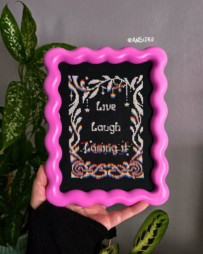 A hand holding a vibrant pink framed craft with Live Laugh Lamington cross-stitch, showing amazing crafts.