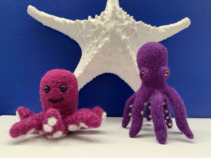 Two small, purple, felted octopus crafts sit in front of a white starfish, against a blue background, displaying amazing crafts.