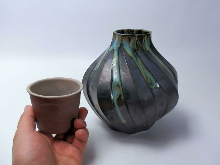 A hand holds a small brown cup next to a larger, dark grey, ribbed vase with blue-green glaze, showcasing amazing crafts.
