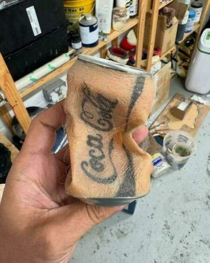 A hand holding a crushed Coca-Cola can that appears to be made of skin with hair and a tattoo, a truly awful idea executed with skill.