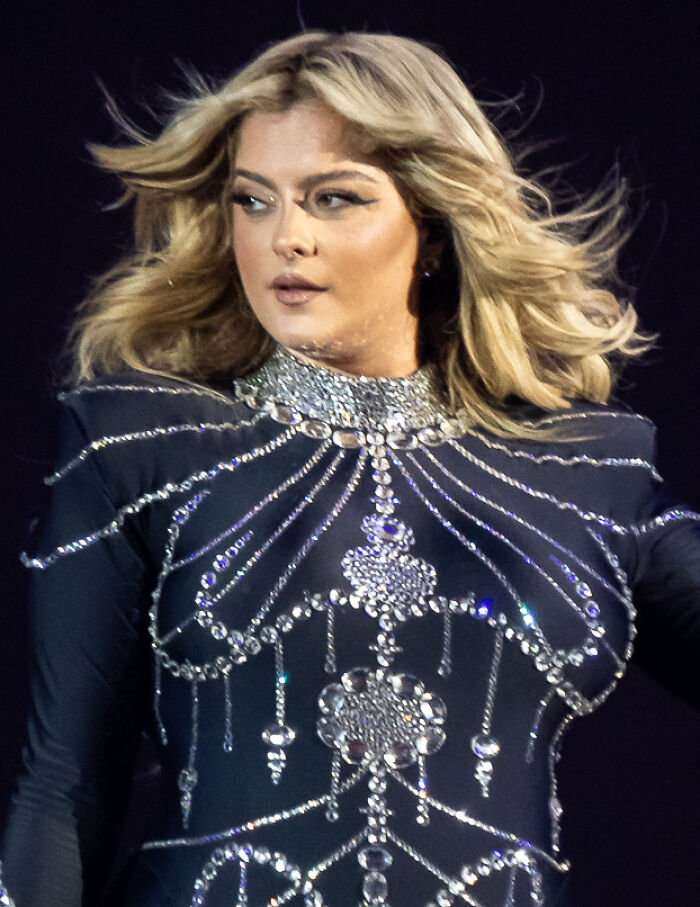 Bebe Rexha on stage, wearing a black long-sleeved top adorned with sparkling silver embellishments, her blonde hair flowing.