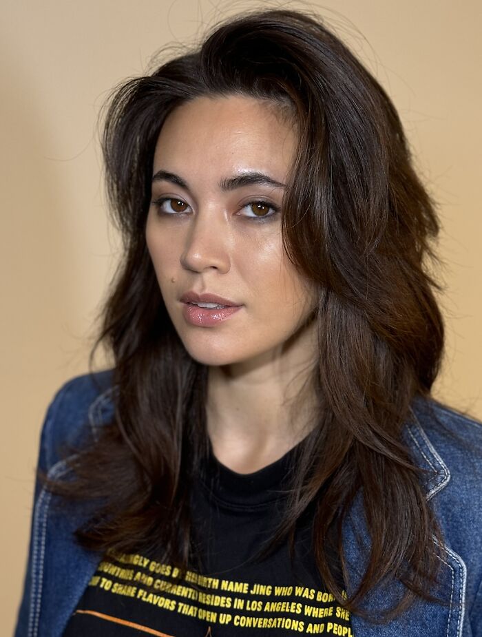 A close-up portrait of Jessica Henwick, an actress with a successful career. Her long dark hair frames her face, enhancing her features. Henwick looks directly at the camera with a confident expression. Her denim jacket adds a touch of casual elegance to her look.