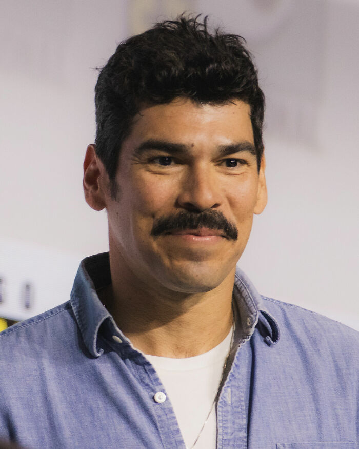 Raúl Castillo smiling, showcasing his career highlights and bio. He is wearing a blue denim shirt over a white tee.
