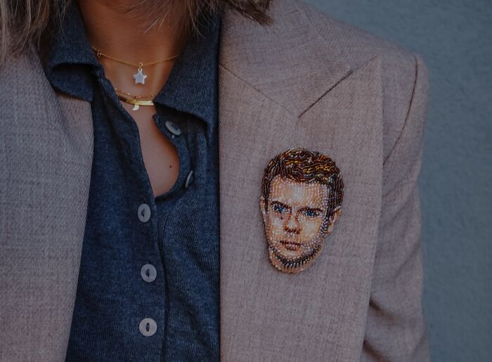 A portrait-style beaded creation by Tiffany Pisani, depicting a male face, pinned as a brooch on a tan blazer.