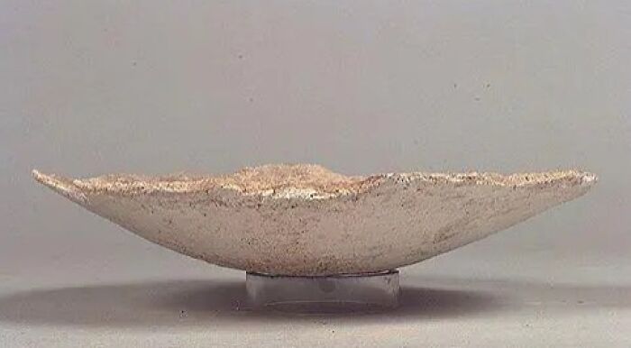 A shallow, textured, light-colored historical artifact bowl on a clear stand, demonstrating its improbable survival.