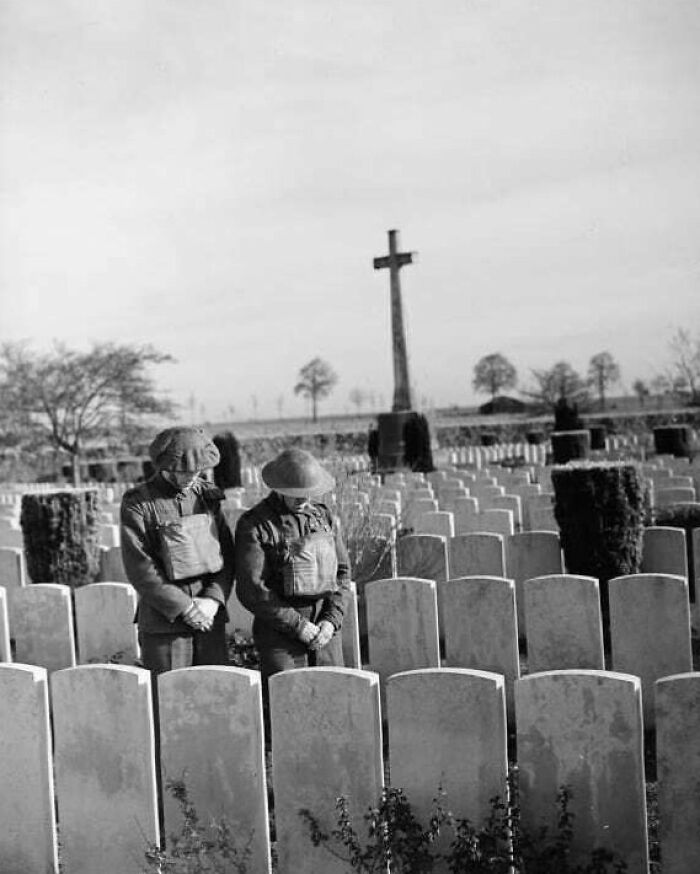 Two soldiers stand reverently in a war cemetery with gravestones and a cross, moments lost to history.