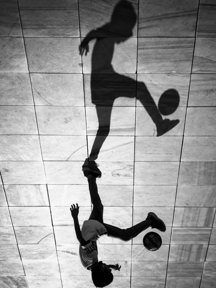 A perfectly timed street photo by Mehmet Bayram shows a boy's reflection playing with a soccer ball, creating an abstract image.