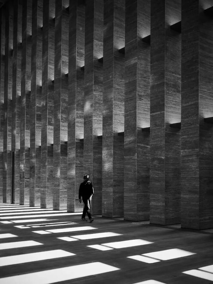 A perfectly timed street photo by Mehmet Bayram captures a person walking through a hall with strong vertical pillars and dramatic light patterns.