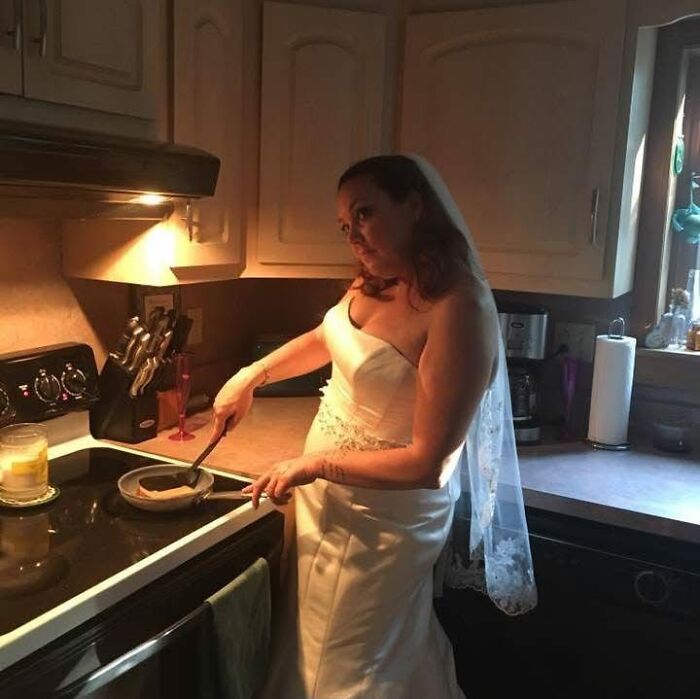 A bride in a white wedding dress fries food in a pan, a funny wedding photo showing her cooking in the kitchen.