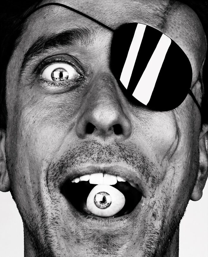 A black and white portrait by Brock Elbank of a man with an eye patch, holding an eyeball in his mouth, an intriguing story.