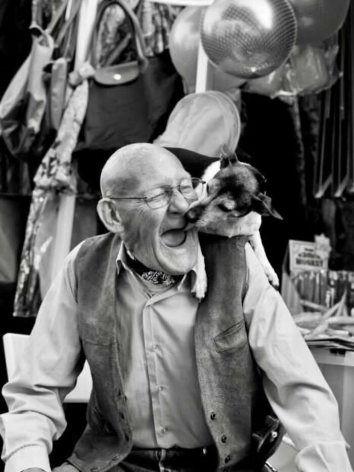 A black and white artistic image shows an older man with glasses laughing as a small dog licks his face.