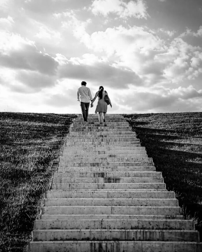 A black and white perfectly timed street photo by Mehmet Bayram captures a couple holding hands, walking up a long staircase.