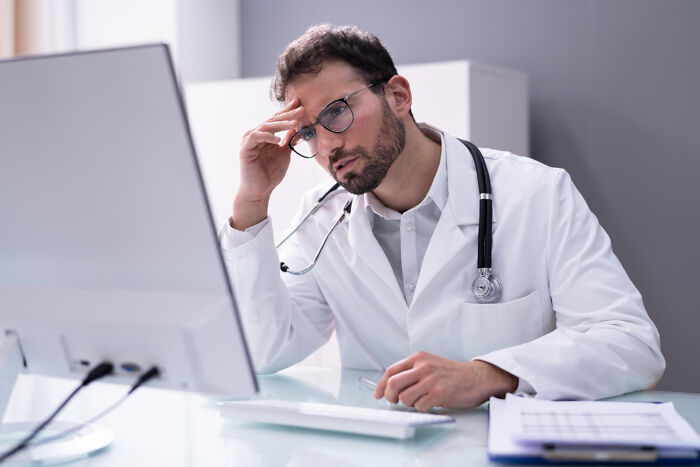 A male doctor looking stressed at a computer, reflecting the difficulty of delivering diagnoses.