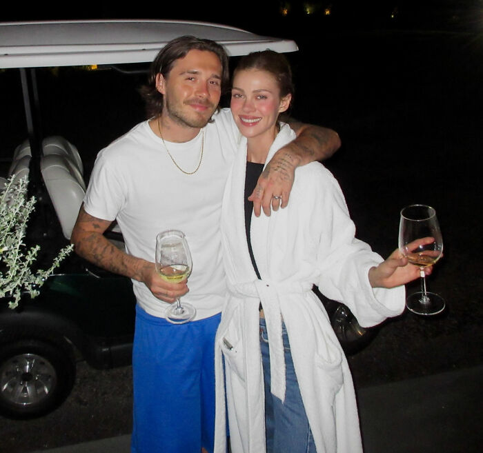 Brooklyn Beckham and a woman, possibly Nicola Peltz, holding wine glasses next to a golf cart.