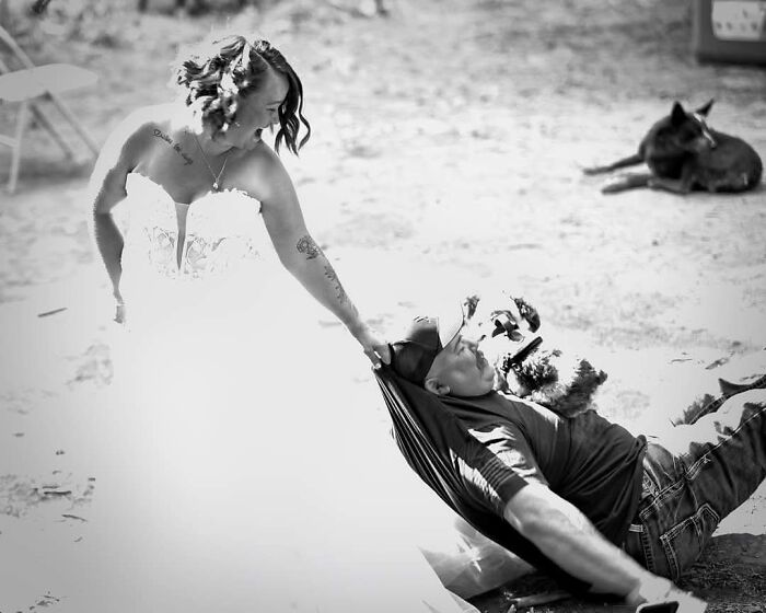 A black and white funny wedding photo of a bride pulling her groom up from a hammock, with a dog in the background.