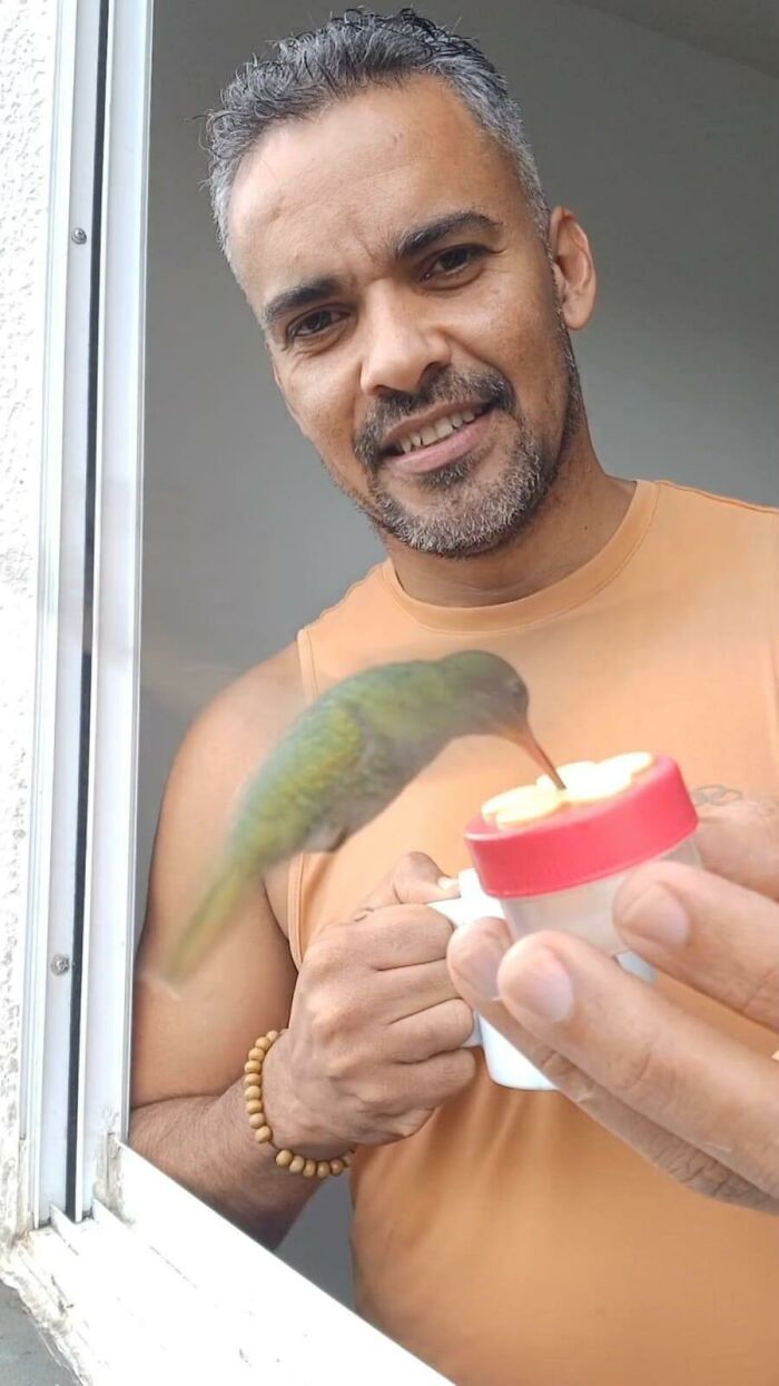 A Brazilian man in an orange tank top smiles as a hummingbird feeds from a small container at his bird restaurant.