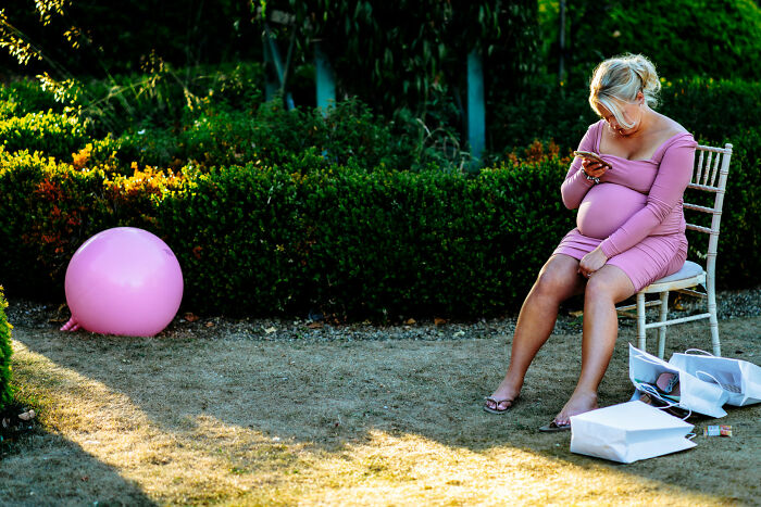 A heavily pregnant woman in a pink dress on a chair next to a large pink balloon, a street photography awards image.